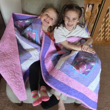 Two smiling girls wrapped in colorful quilts.
